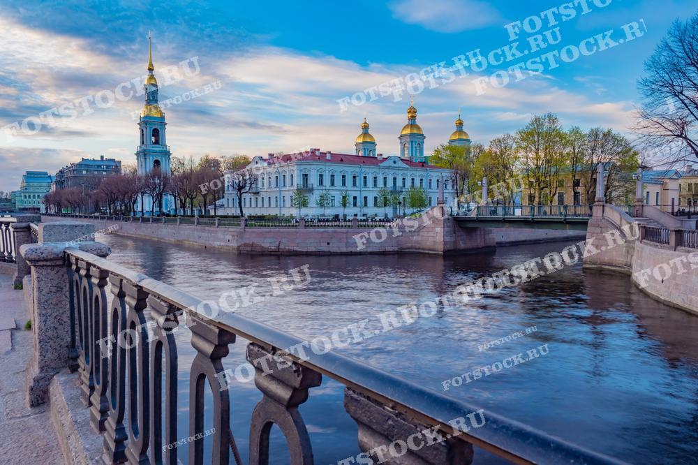 На фотографии изображен Никольский морской собор и канал Грибоедова в Санкт-Петербурге. Весенний день и спокойная вода создают атмосферу умиротворения. Фото может быть использовано для туристических буклетов, веб-сайтов о Санкт-Петербурге, а также для различных личных и коммерческих проектов, связанных с культурой и архитектурой города.
